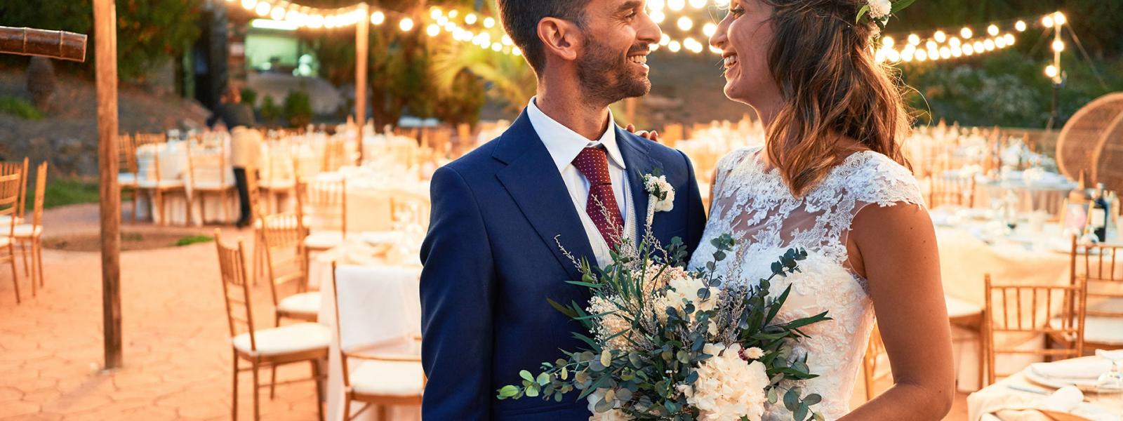 Wedding couple in a courtyard setting with tables and chairs in the background and lights going around the back of them in a circular shape